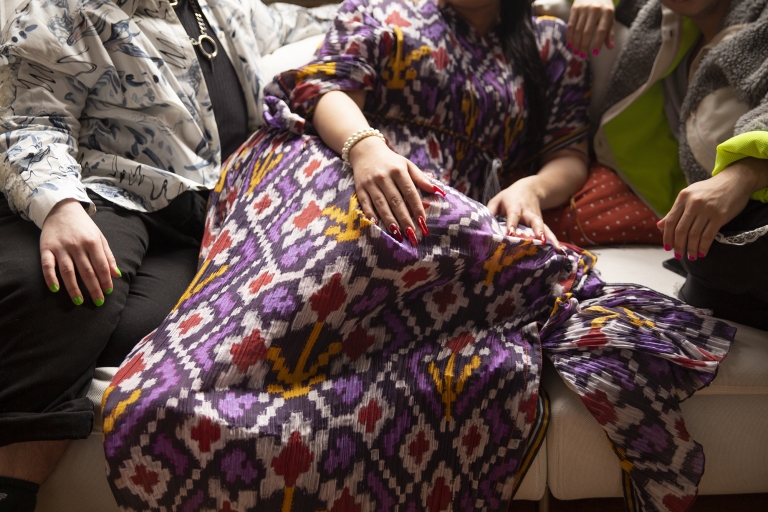 Three people cuddle together on a couch but only their shoulders and down are seen. The person in the middle is wearing a colorful pattern with long red nails.