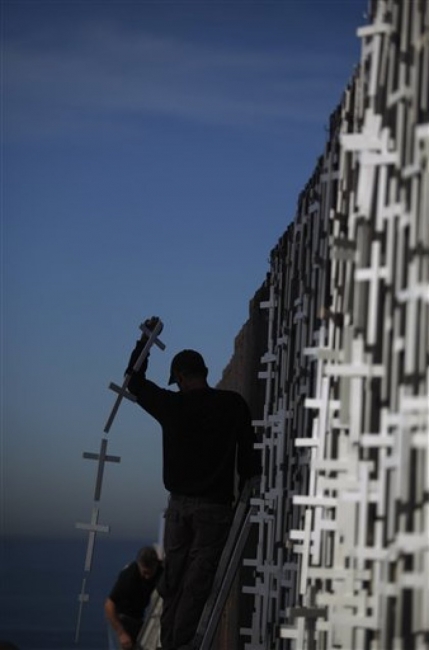 A person stands on a ladder against a large wall of small crosses. The person is holding up a string of crosses.
