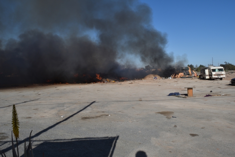 An asphalt lot is filled with fire and smoke. An RV is seen off to the side.