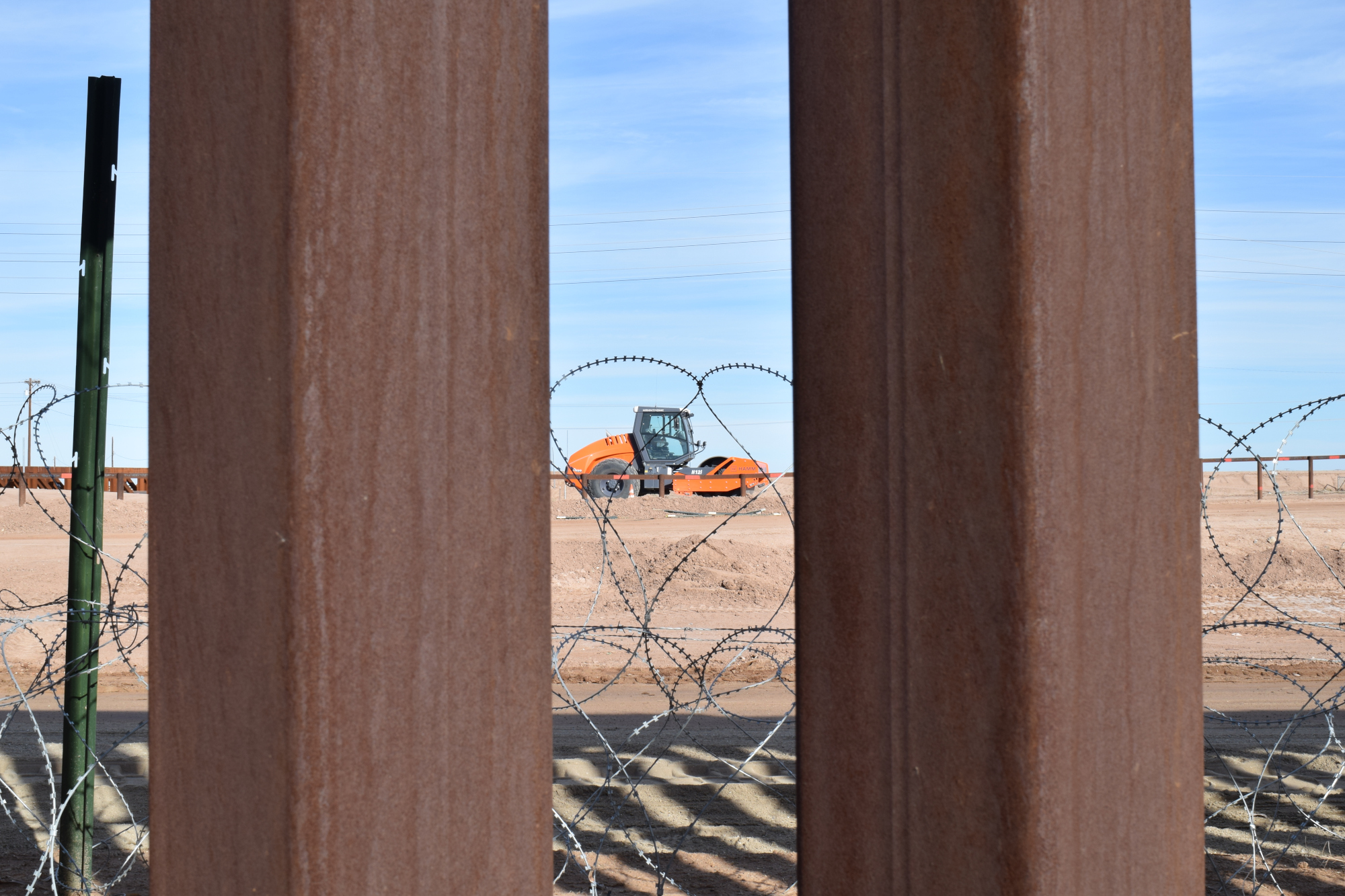 The scene is framed by rusted steel beams. Between the beams there are coils of barbed wire and a large construction machine.