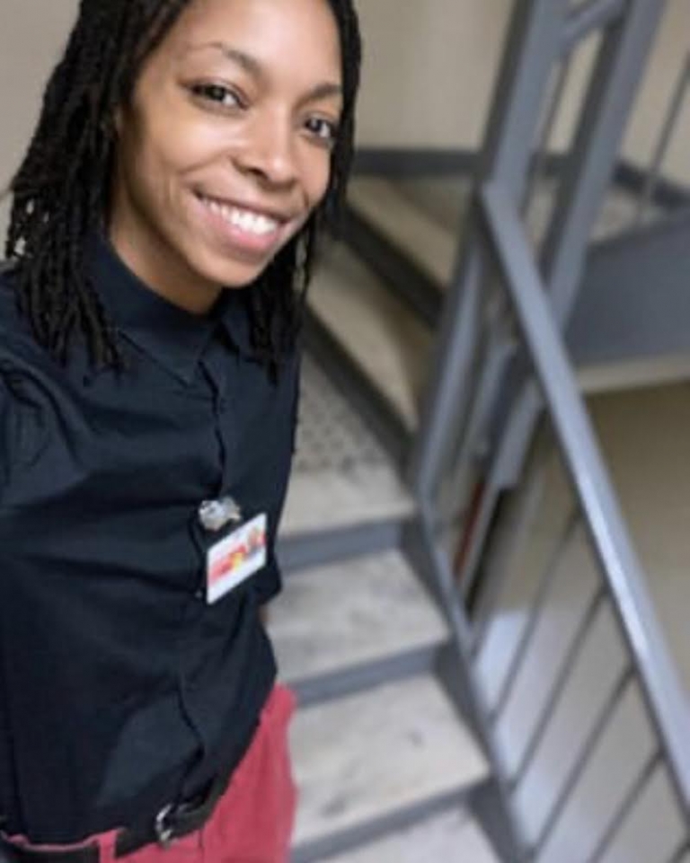 A Black person with long hair wearing a black shirt and red pants poses in a stairwell