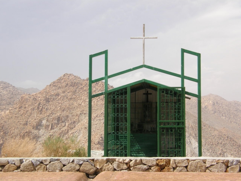 A green building with a cross on top of it with mountains in the back.
