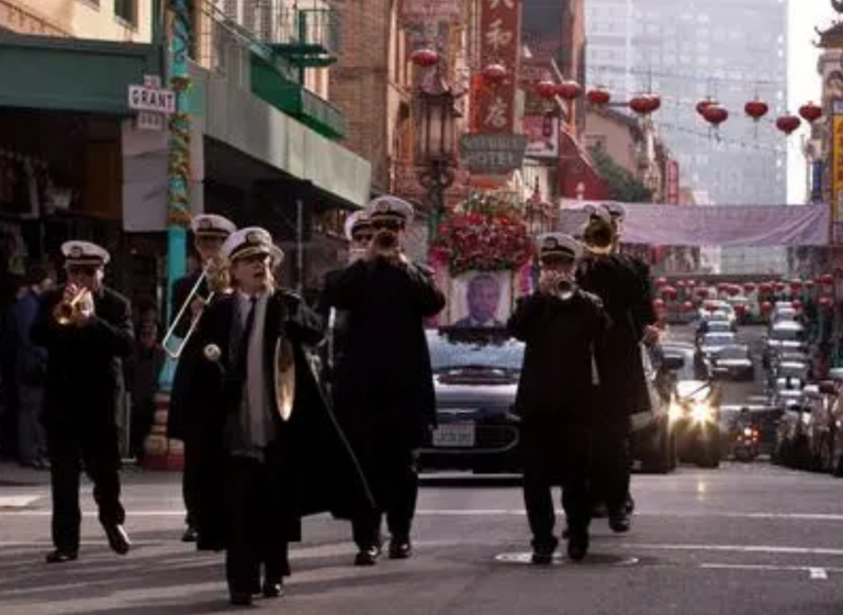 A group of people walk down the street playing instruments. Behind them follows a casket.
