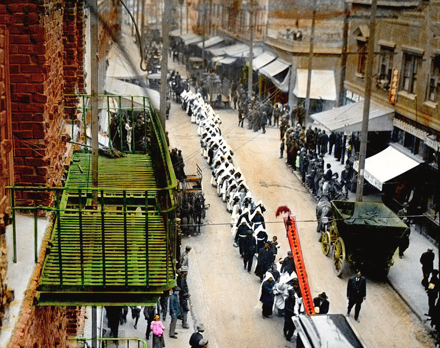 Looking down on a dirt street there is a procession of people dressed in black with white robes. People are carrying red banners and towards the back there is a casket.