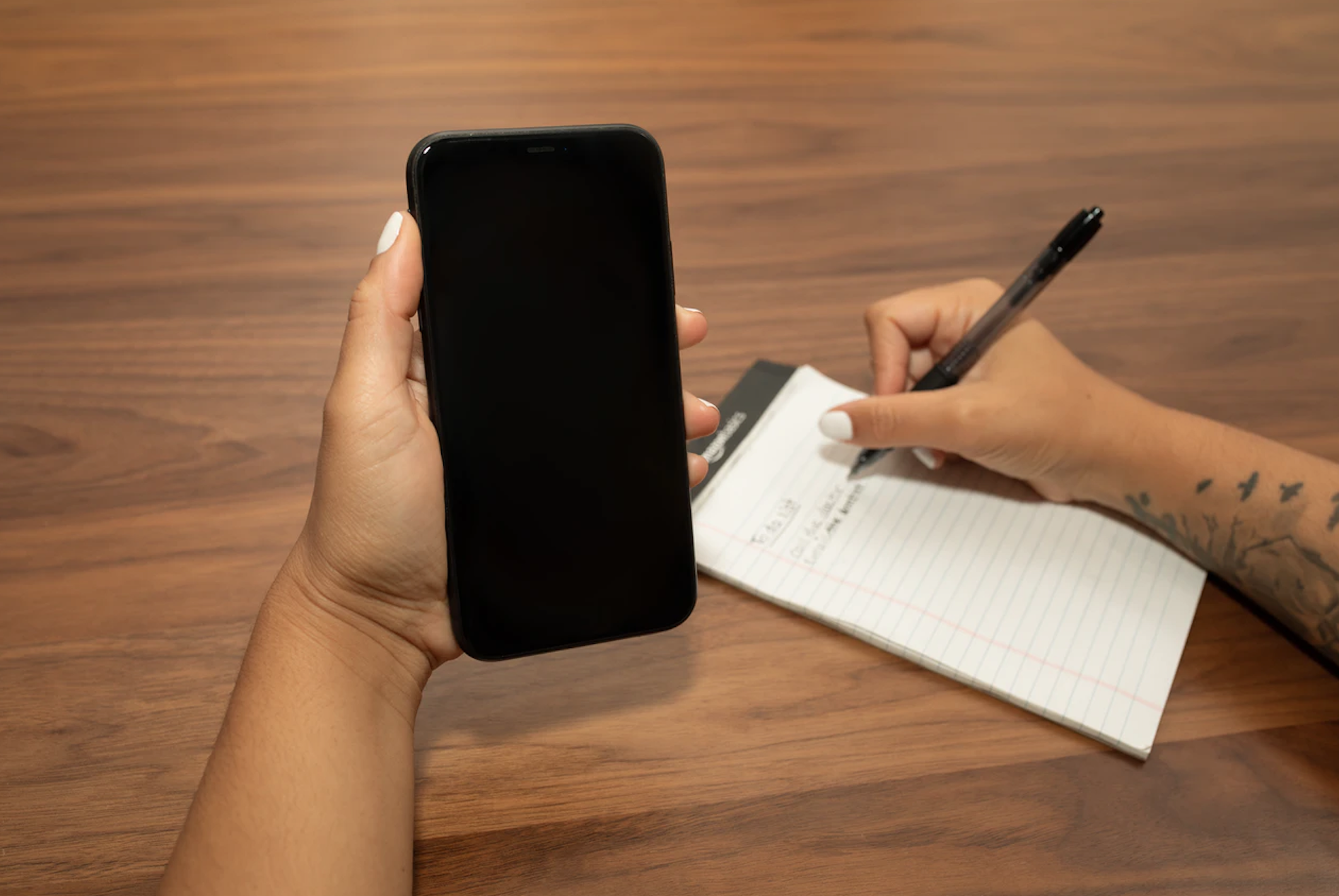 Hands holding a phone while another hand is writing on a notepad with a pen.