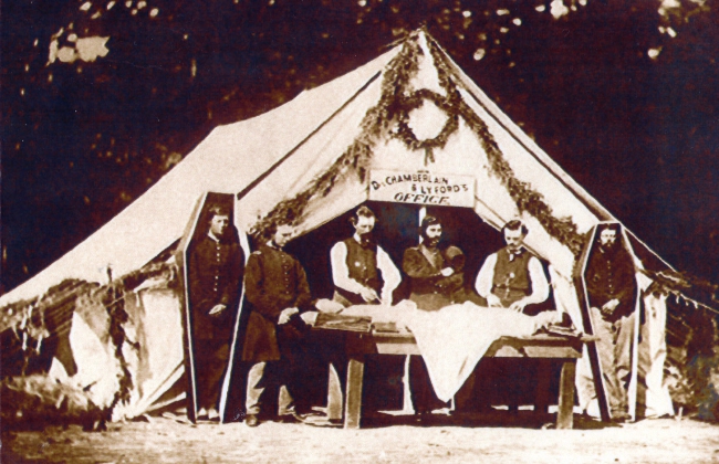 Sepia toned photo from the Civil War Era of four men standing between a large white tent and a wooden table with a white fabric wrapped corpse on top of it. On either sides of the men are two corpses standing in their coffins. There is a sign above them that reads, 