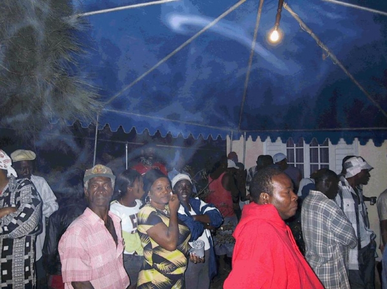 Jamaican men and women are gathered outside together under a tent awning.