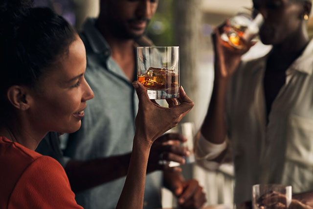 A woman gazing into a glass filled with a brown liquid and ice cubes. She is surrounded by men drinking from their own glasses.