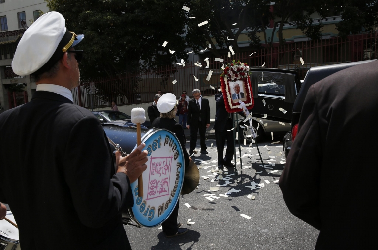 A person in uniform beats on a drum as they walk down a street filled with confetti flying.