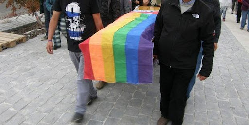 A group of pallbearers carry a casket covered in a rainbow flag down a street