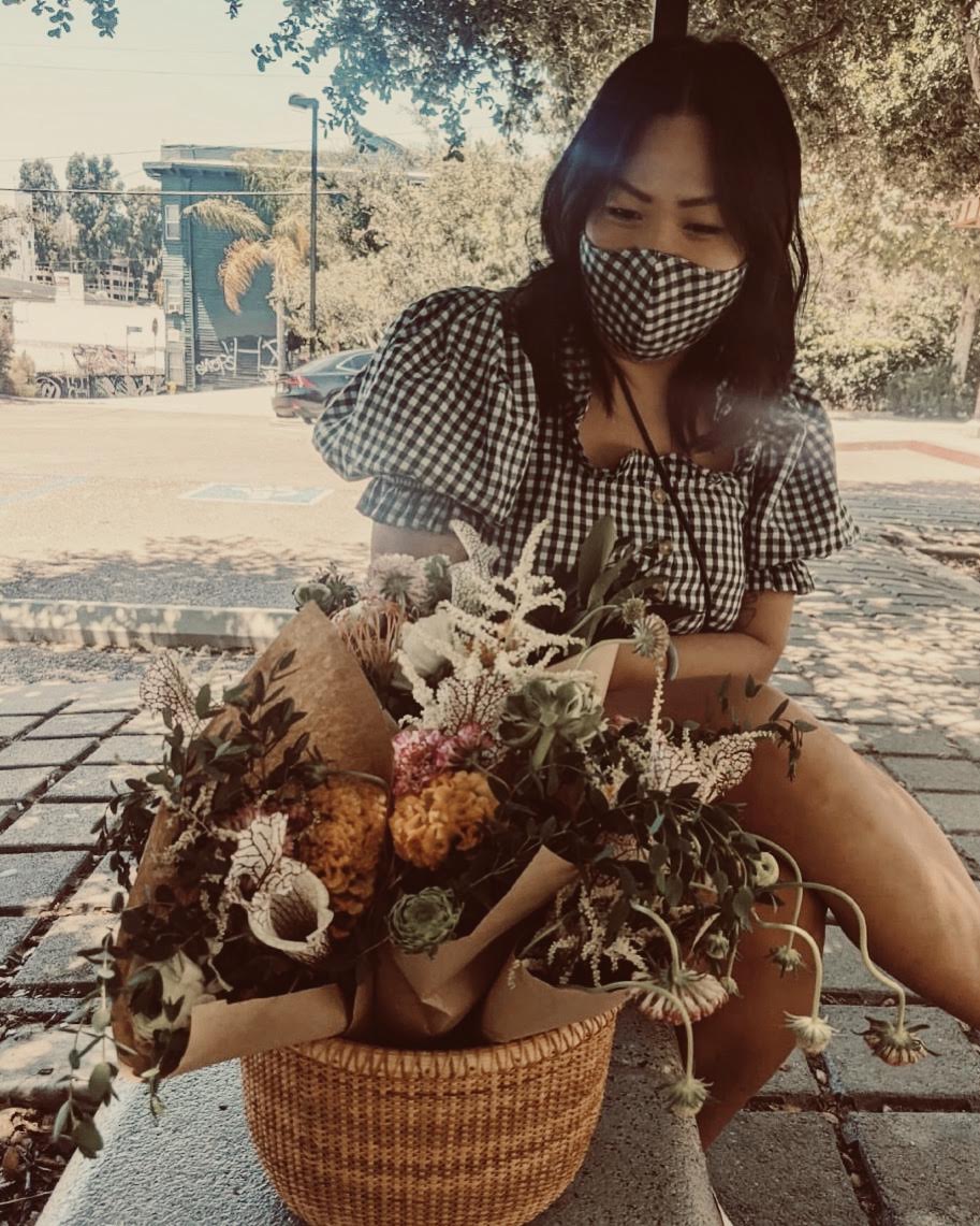 A medium skin toned woman wearing a mask and black and white dress, sits alongside a basket of flowers