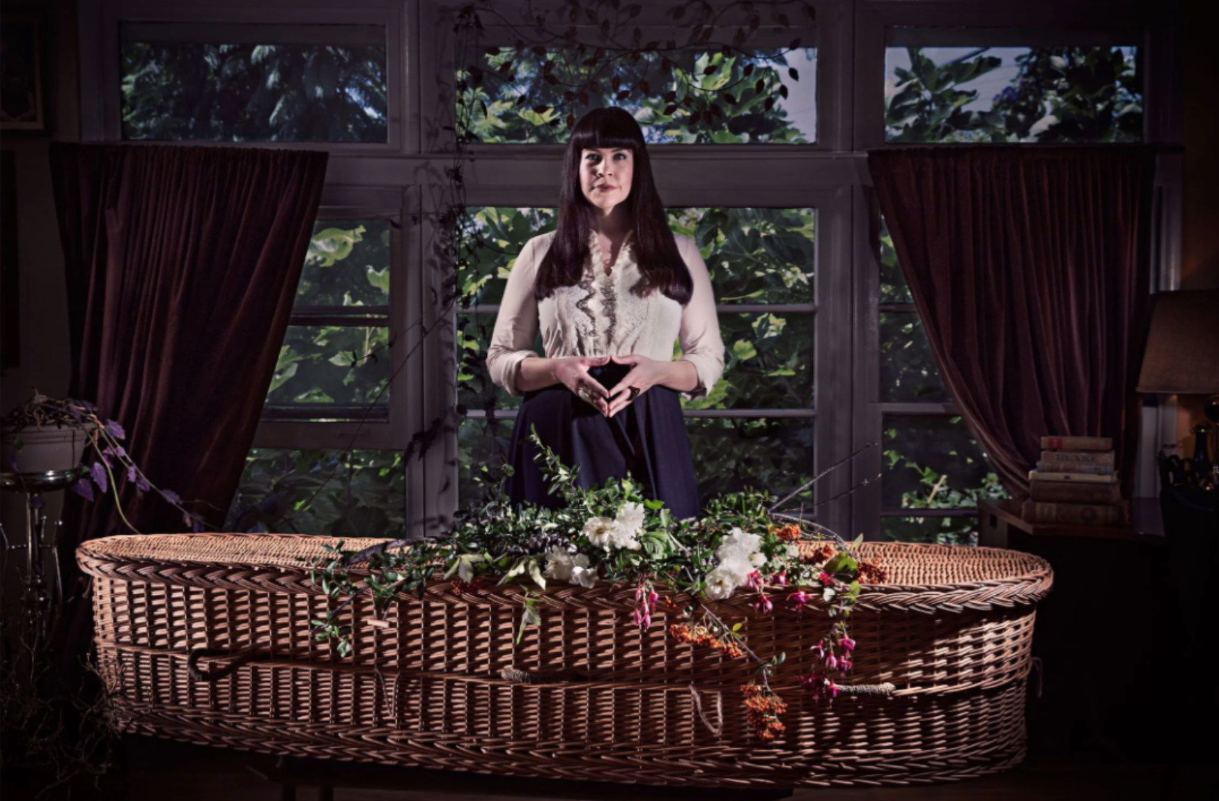 A dark-haired woman in a ruffled blouse with hands together over a woven basket casket covered in flowers. This is the Order of the Good Death founder, Caitlin Doughty
