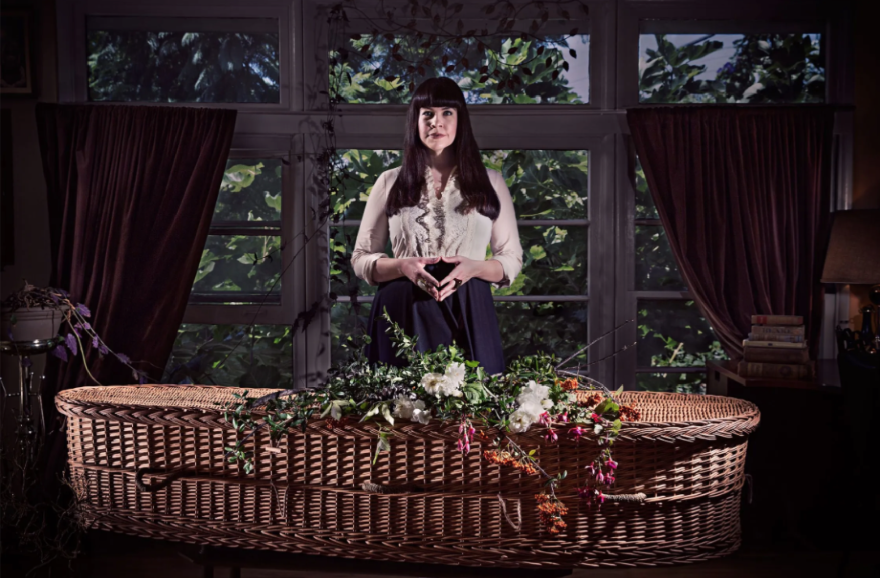 A light skinned woman with dark hair stands behind a wicker casket in a living room