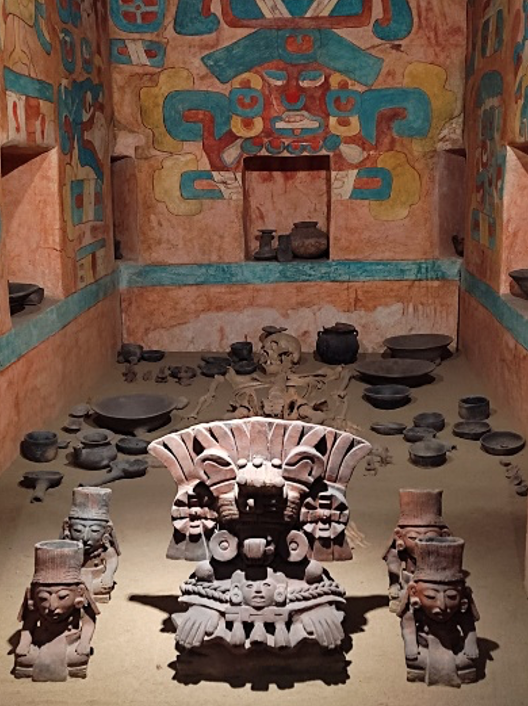Recreation of a pre-hispanic tomb. On the floor are skeletal remains and various clay artifacts. The surrounding walls are terracotta color with colorful painting over it.