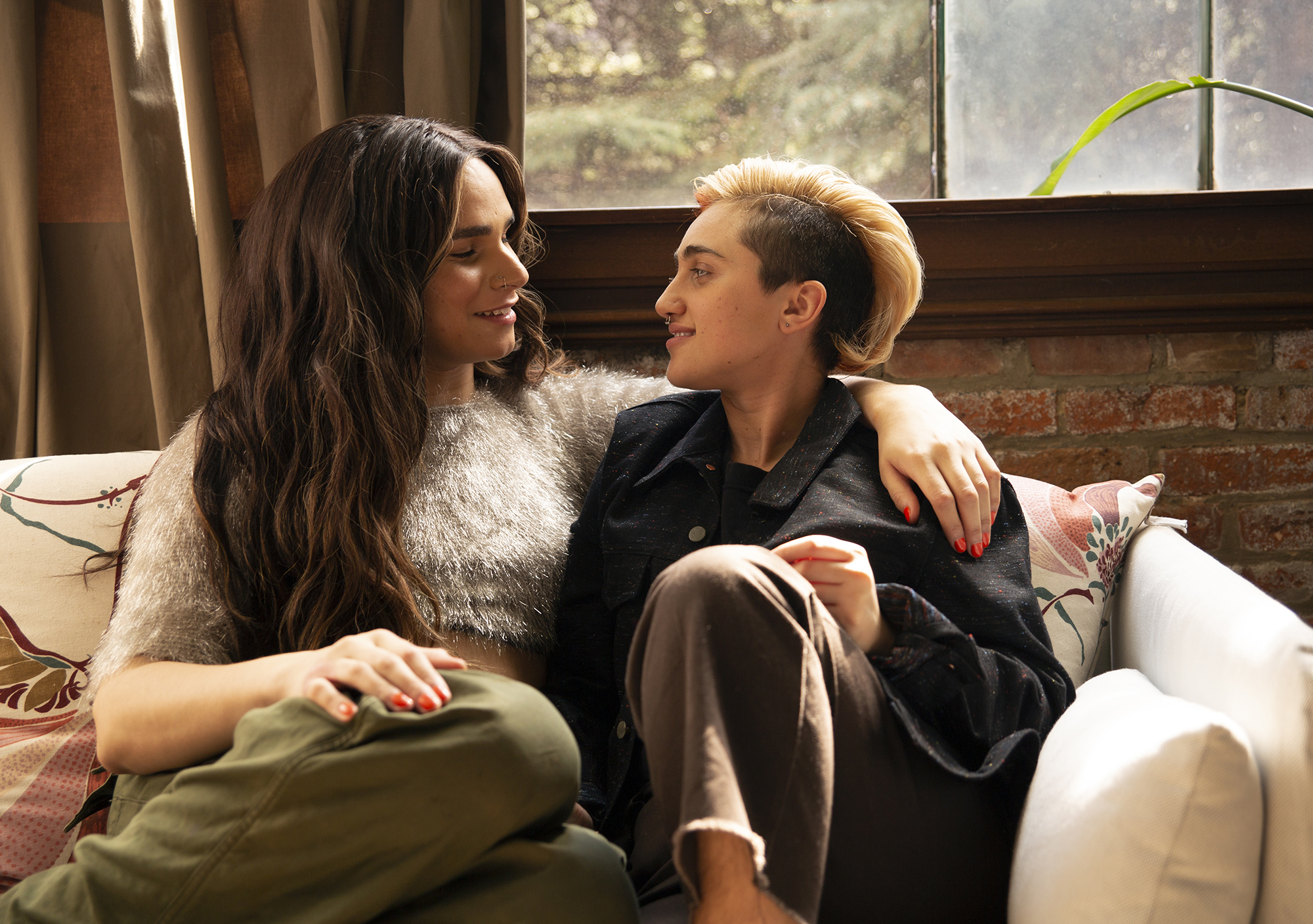 Photograph of queer couple sitting on couch looking at each other.