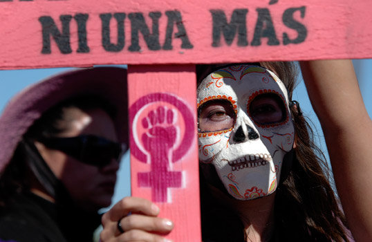 Photo of a woman with her face painted as a skull holding a sign that reads 
