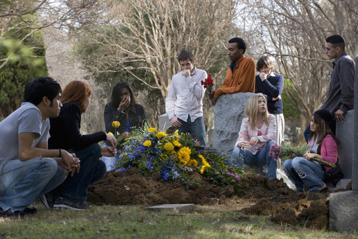 Photo of a group of people grieving around a fresh grave covered in flowers.