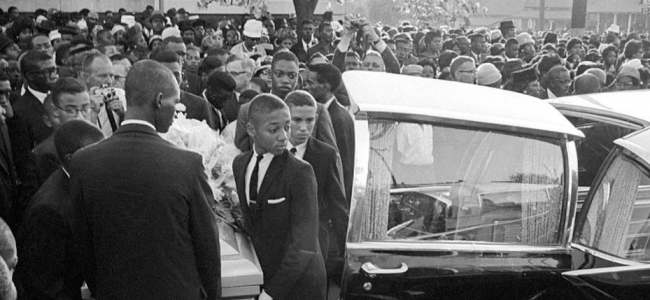 Black and White photo of young Black men carrying a casket into a funeral hearse.