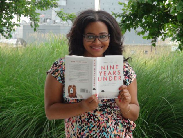 A dark skinned woman wearing glasses is smiling, she stands in front of tall grass holding up a book. The book cover reads Nine Years Under in red lettering