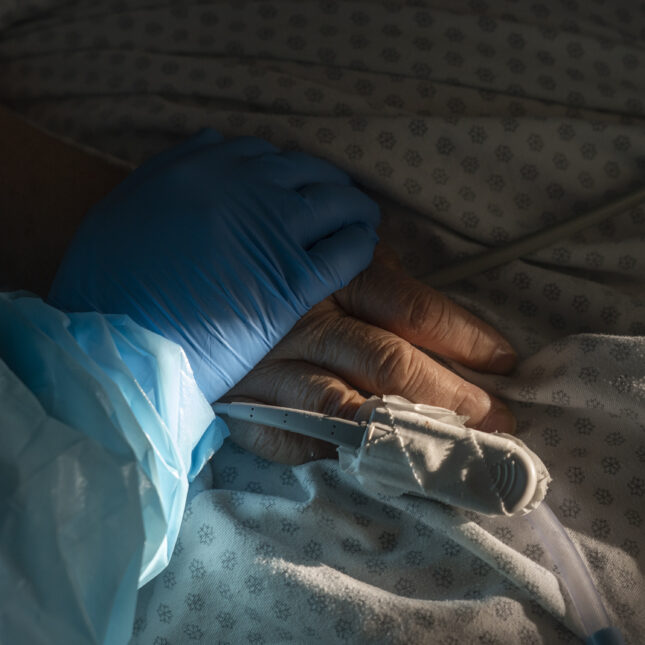 Close up of a bare hand resting on top of a person wearing a hospital gown that belongs to a patient. A blue gloved hand belonging to a health care provider holds the patients hand.