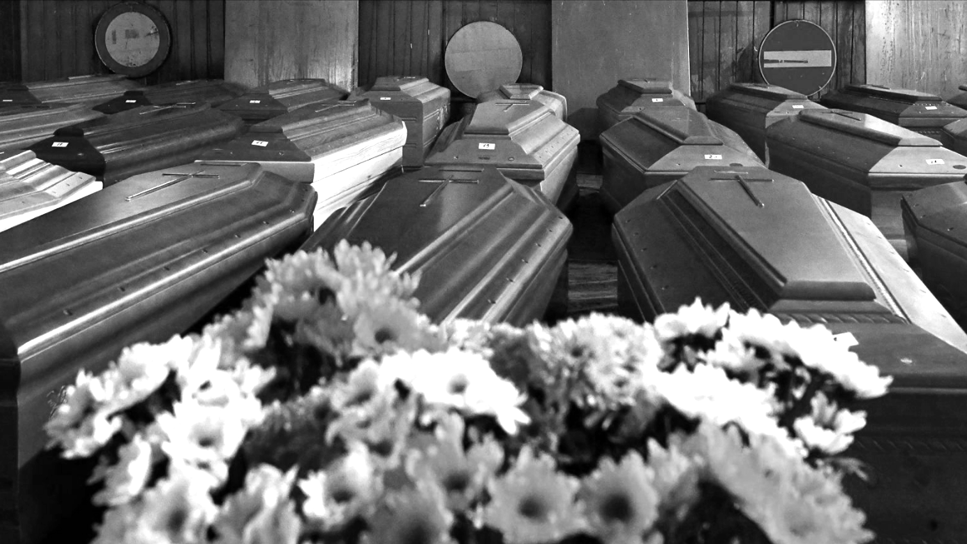 A black and white photo of a room packed with caskets, with white flowers in the foreground.