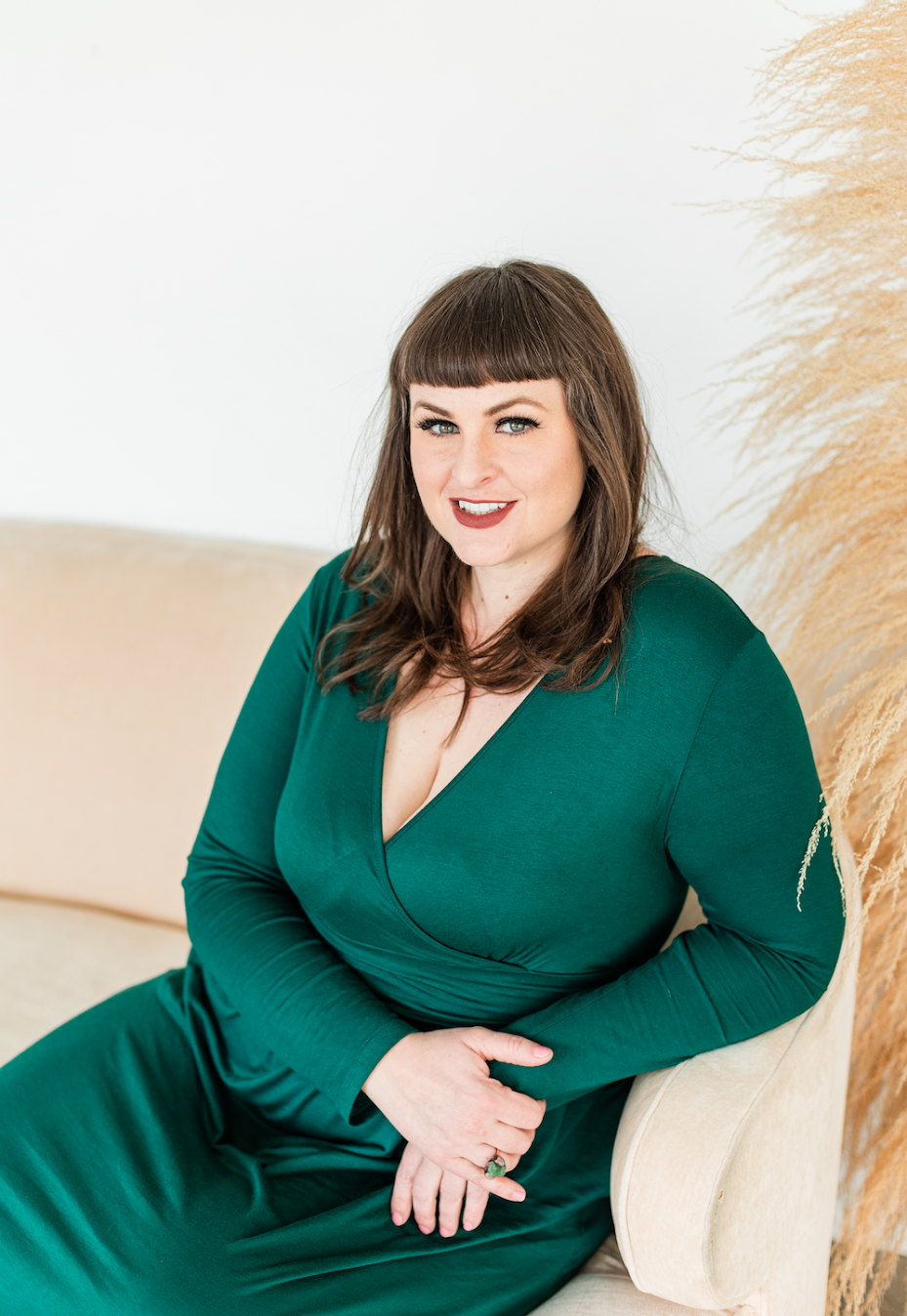 A woman with dark hair and bangs, sits on a couch wearing a green dress