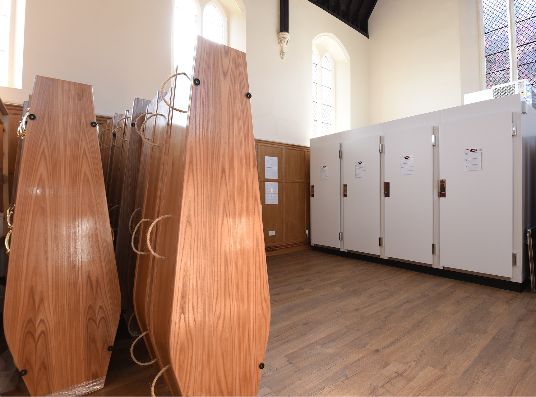 A room filled with natural light, on the left are stacks of wooden coffins standing upright, on the right are three mortuary freezers