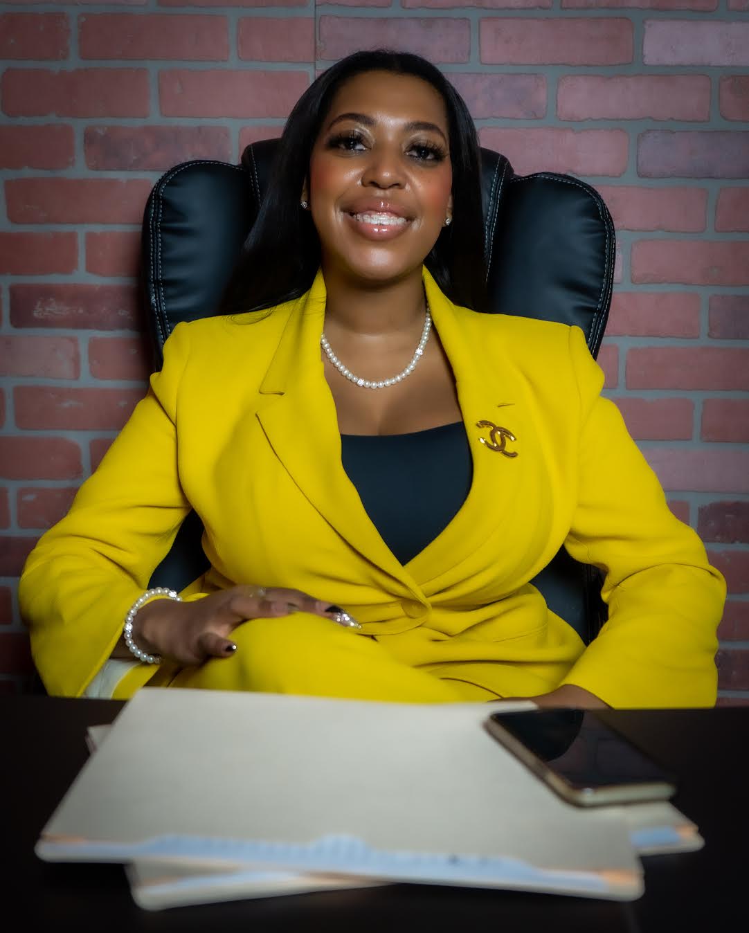 A Black woman wearing a bright yellow blazer smiles while sitting at a desk.