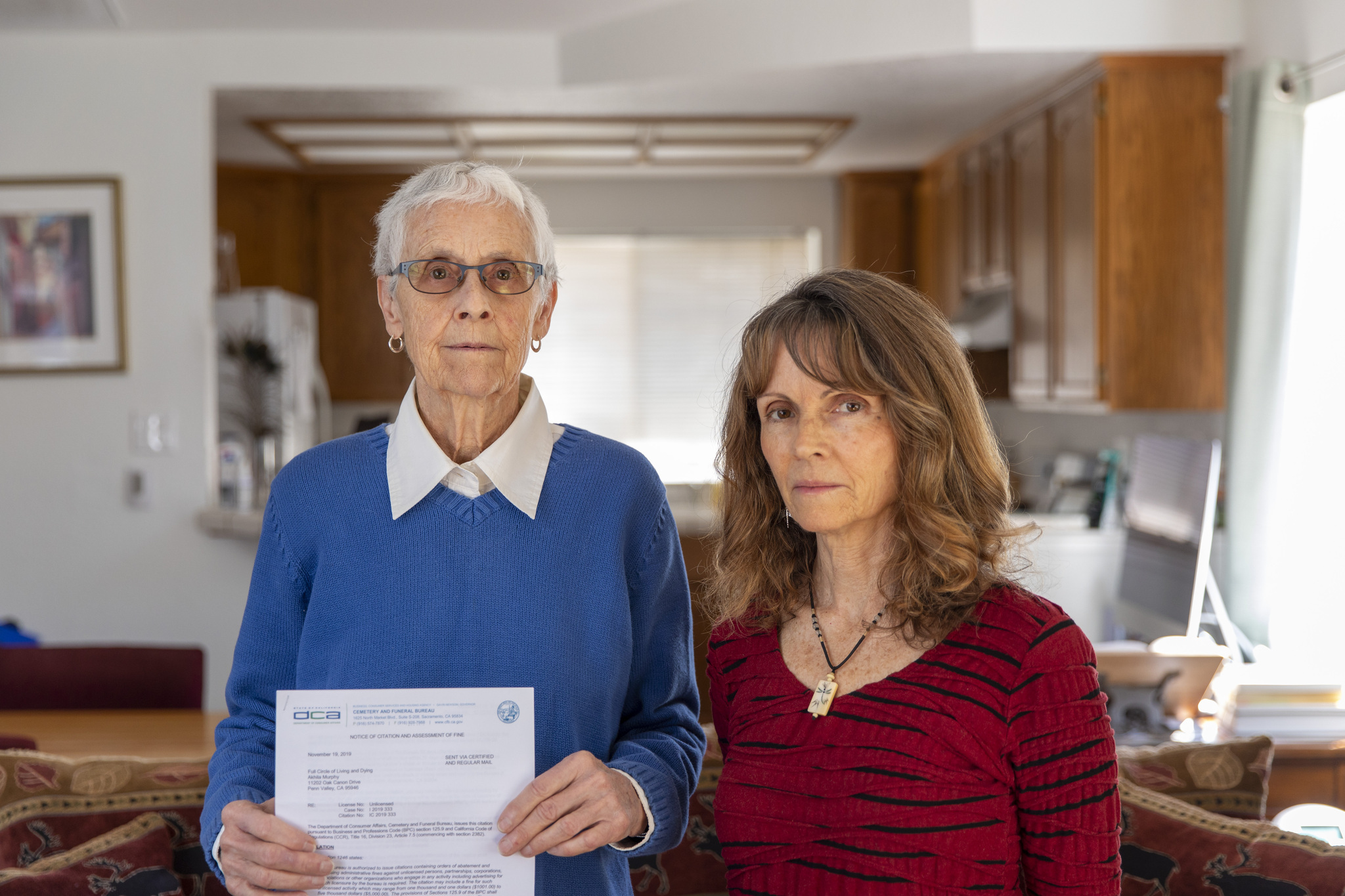 Two women stand side by side, looking at the camera with sober expressions. One of them is wearing a blue sweater and holds a paper, the other wears a red sweater