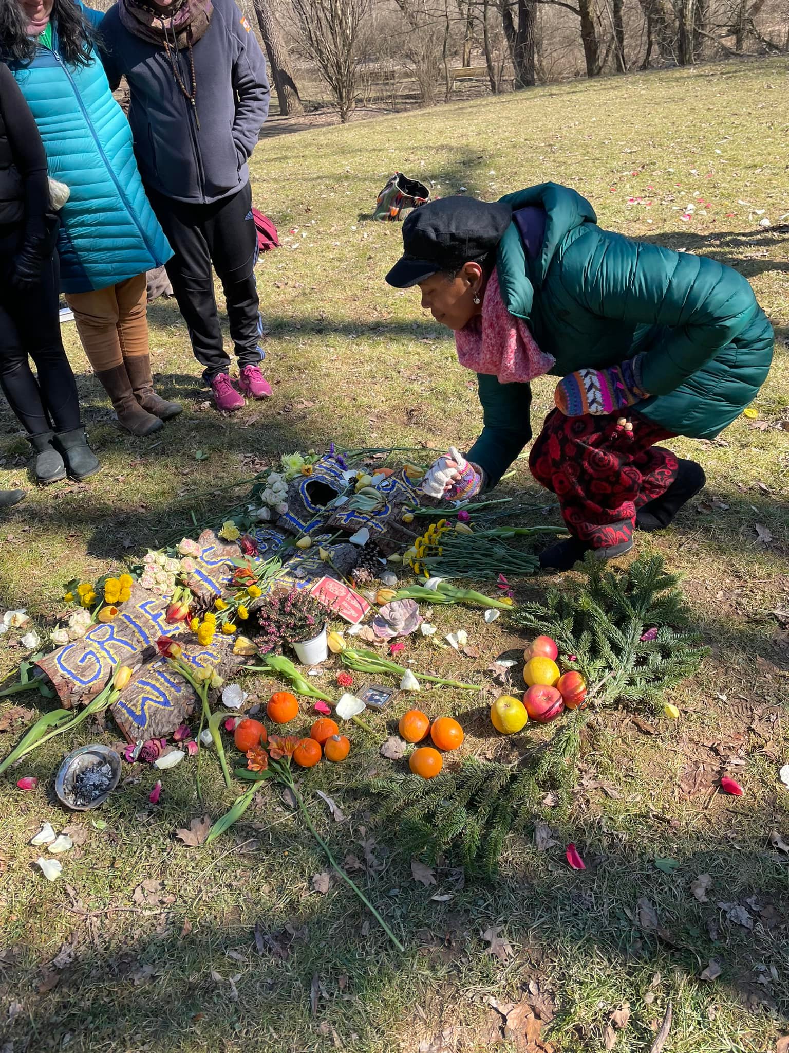 A Black woman brands over an arrangement of flowers and objects, as several prone stand nearby to observe.