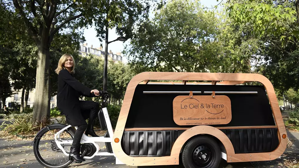 A blonde woman is outside in a park-like area, posing with a bicycle that has a wooden carriage attachment large enough to fit an adult size casket