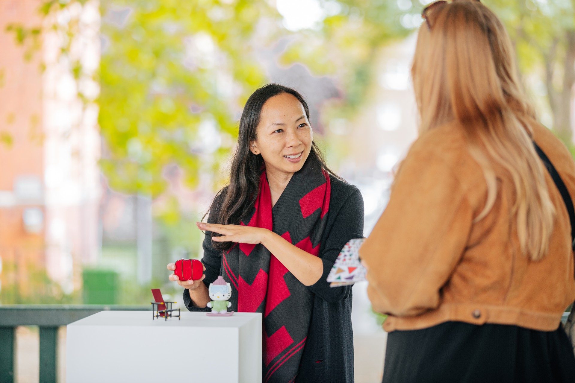 A woman standing behind a pedestal with three objects and restoring toward them while talking to another woman whose back is to the camera