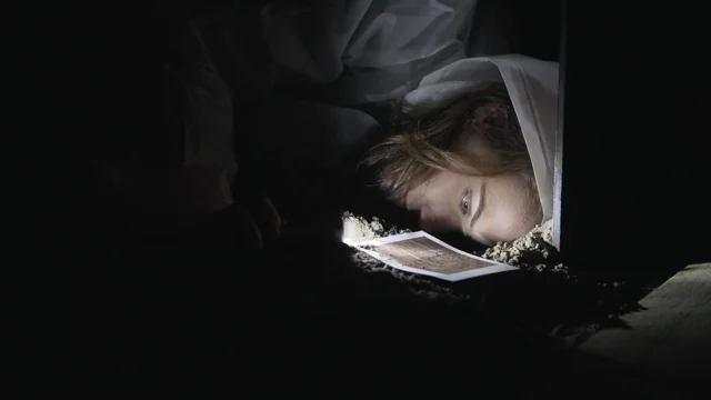 In the darkness, a person lays their ear to the dirt ground. They are wearing a translucent poncho with the hood pulled up over their hair. A light from a flashlight illuminates their face.