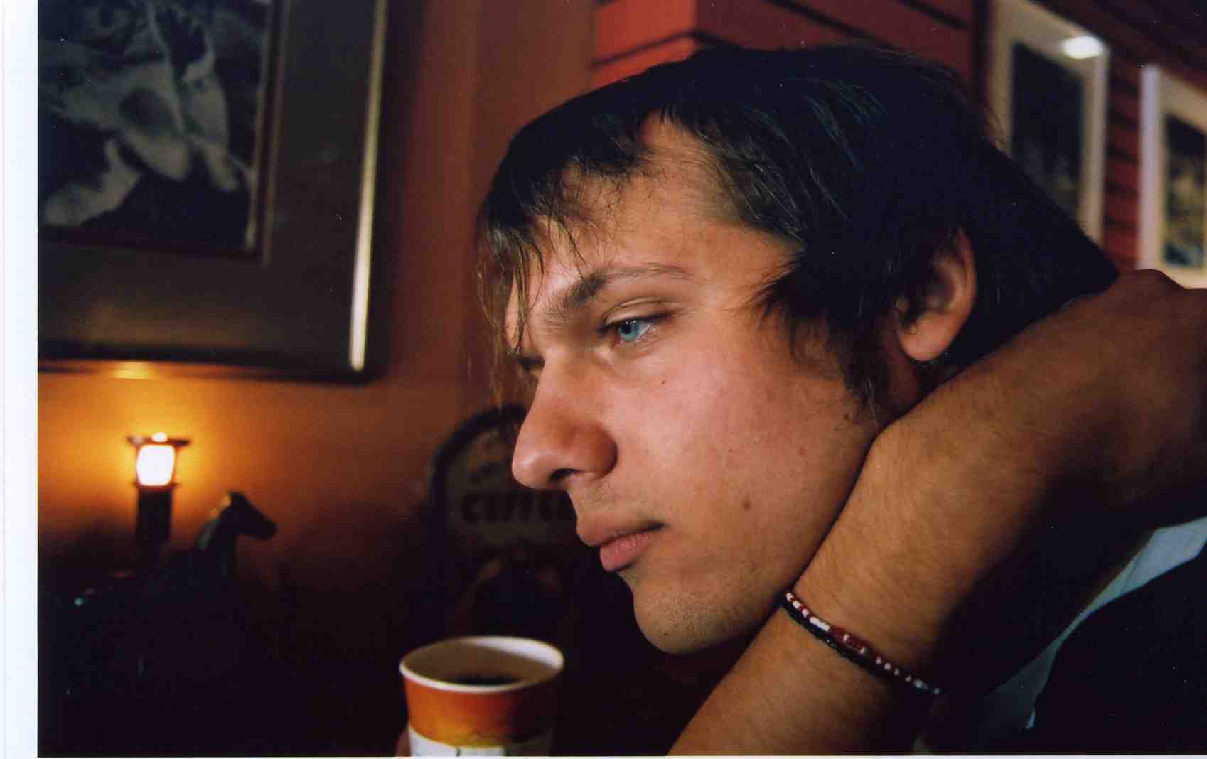 Side or profile view of a man sitting in a room with low light, there is a cup sitting near him. On the walls around him hang various pieces of framed art one of them is illuminated by a candle beneath it.