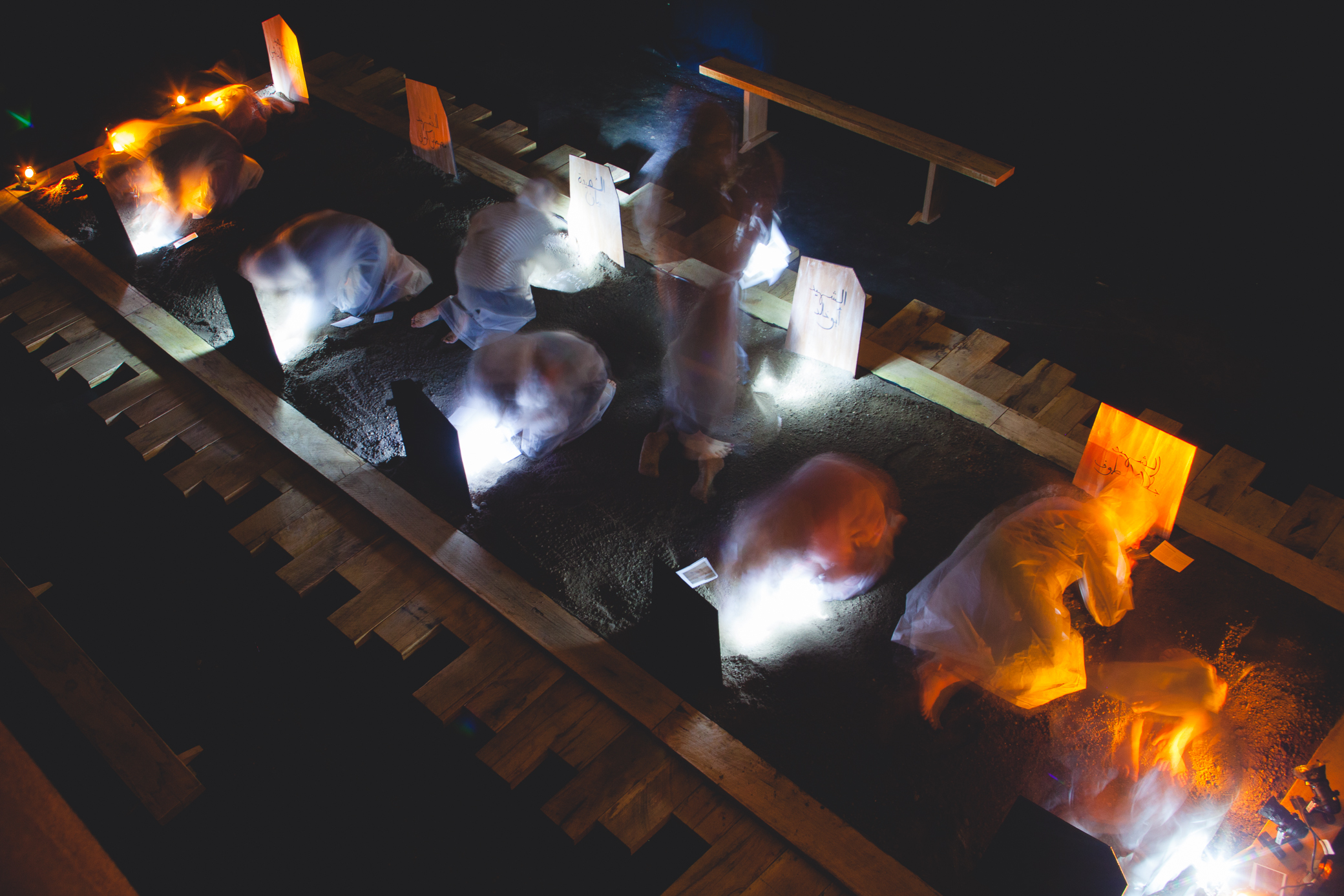 A gallery is transformed into a graveyard. In the middle of the room people wearing translucent rain ponchos, lie down in the dirt. Flashlights sit on the ground nearby each of them. The room is dark.