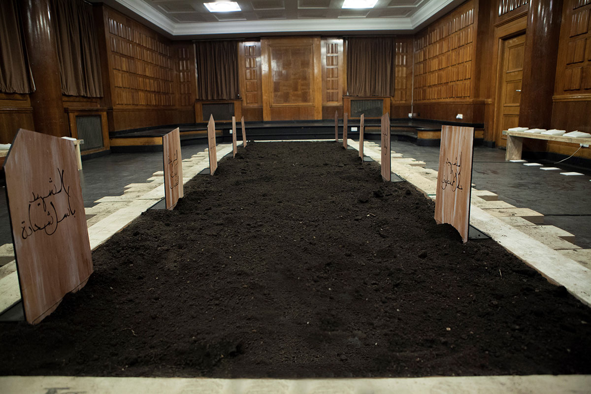 A gallery room with wooden walls and floors. In the center dirt lays on the ground, surrounded by grave markers.