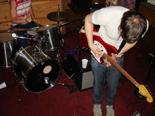 Chris leaning into playing guitar, a drum set sits behind him.