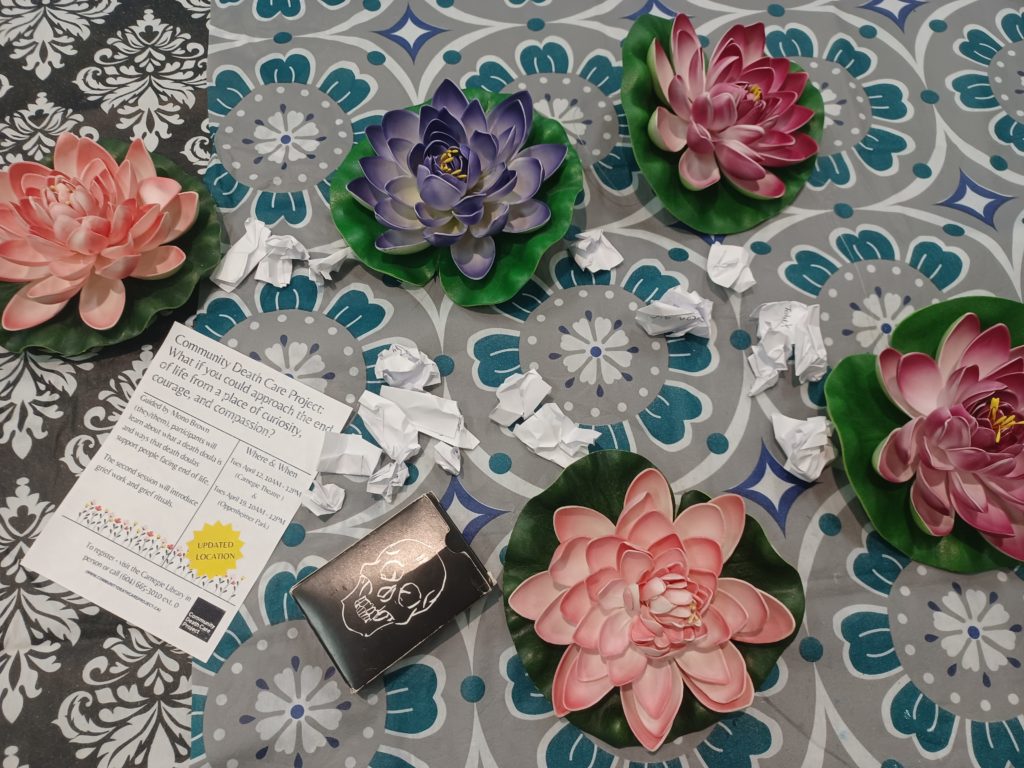 Lotus flowers sitting on top of a blue and white patterned fabric. A flyer with printed information sits alongside a postcard featuring an imaging of a skull