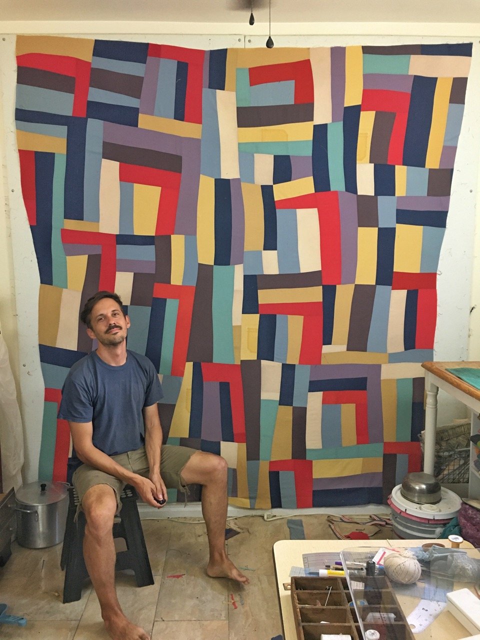 Quilt artist, Zak Foster sits in front of his hanging burial quilt. The colors are a combination of red, blue, yellow, and soft earthy tones.