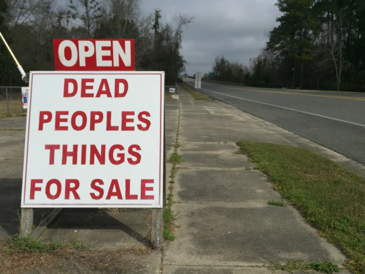A white sandwich board style sign stands on a sidewalk near an empty street. In red lettering the sign reads Open, Dead people's things for sale