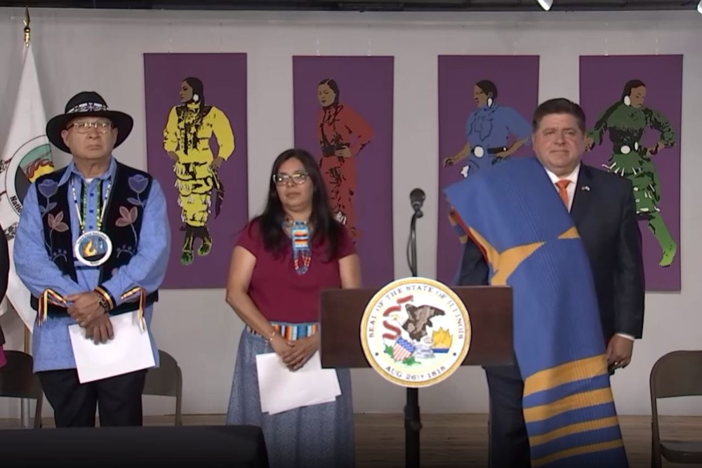 Photo of three people standing behind a podium with a state of Illinois seal on it. On the wall behind them are four art portraits of i indigenous women dancing in regalia
