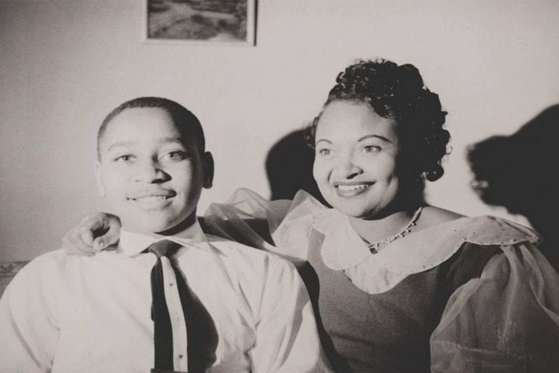 Black and white photograph of Emitt Till, smiling, as he sits next to his mother, Mamie, with her arm around him.