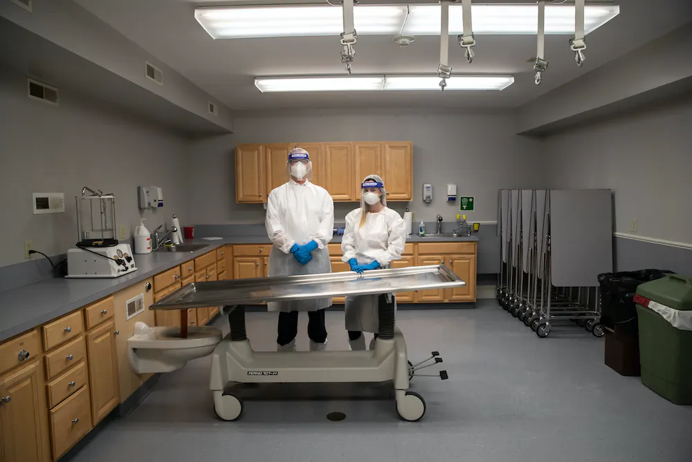 Two people stand in ppe in an embalming prep room