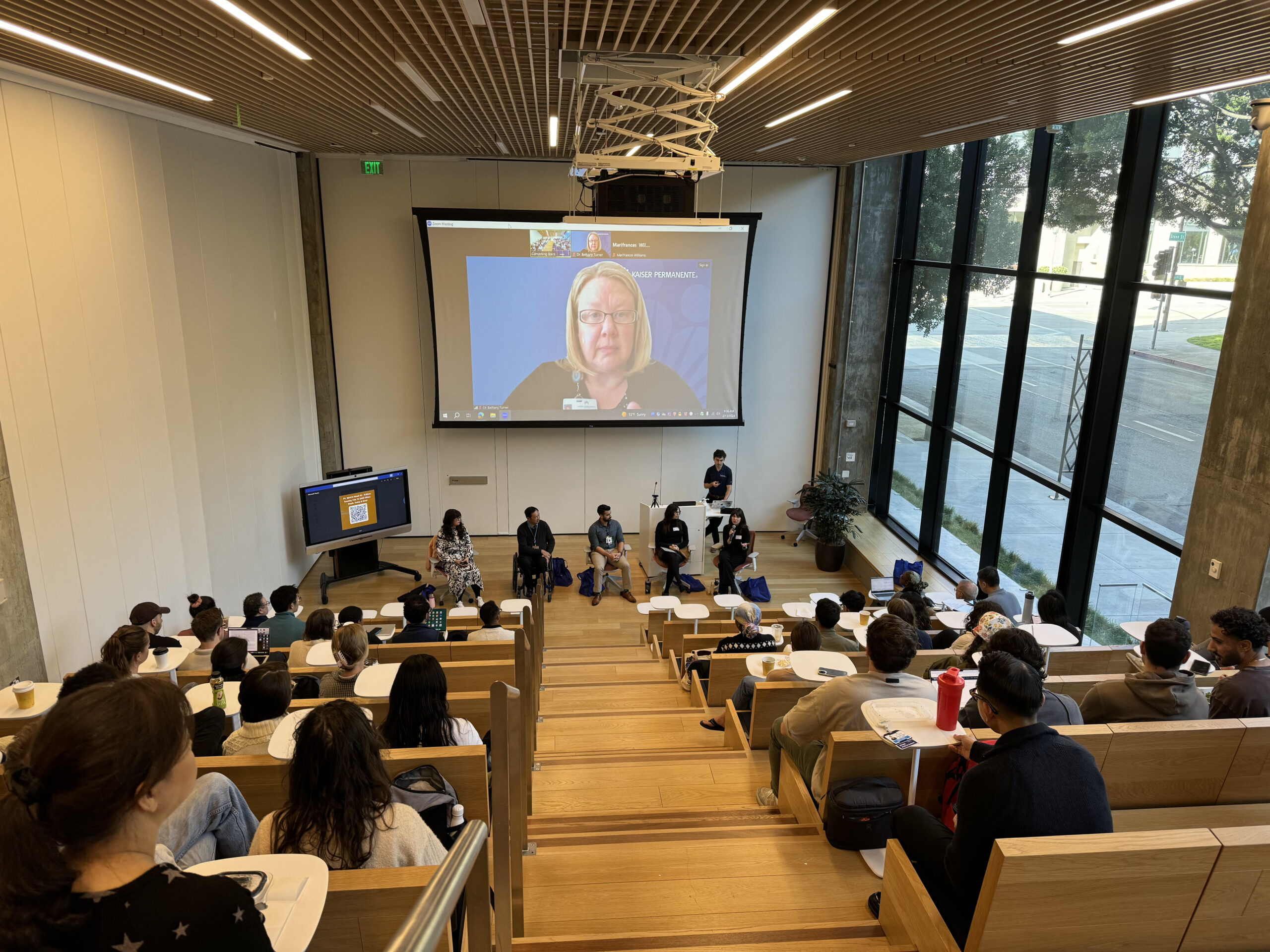 wide shot photograph of a large stadium style modern classroom. Students sit while a panel of instructors sit in front of a large screen.
