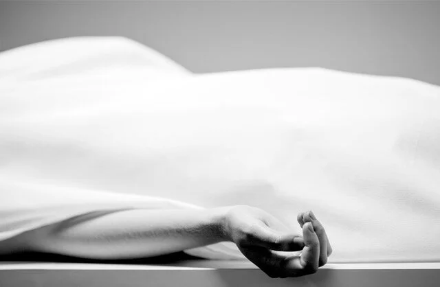 Black and white photograph of a corpse covered with a sheet. A single arm, uncovered, is visible