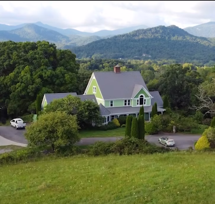 A large home painted light green with a grey roof situated in a grassy hilled area