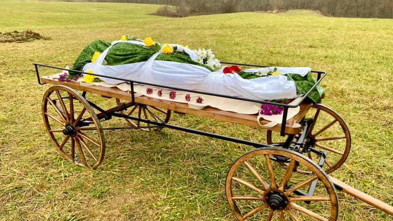 photo of a wooden cart with wheels in an open grassy field. On the cart is a shrouded body adorned with flowers