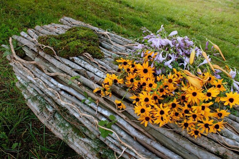 Photo of a beautiful coffin fashioned out of tree branches. The coffin lays on the grass and has yellow and purple flowers on top of it